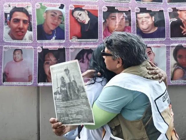 Dos mujeres que buscan a un familiar reportado como no localizado se abrazan frente a una lona con fotografías de personas desaparecidas. Foto: Cortesía.